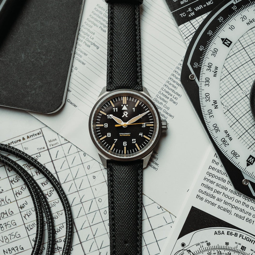 Black wristwatch with leather strap on a white surface with technical drawings and documents.