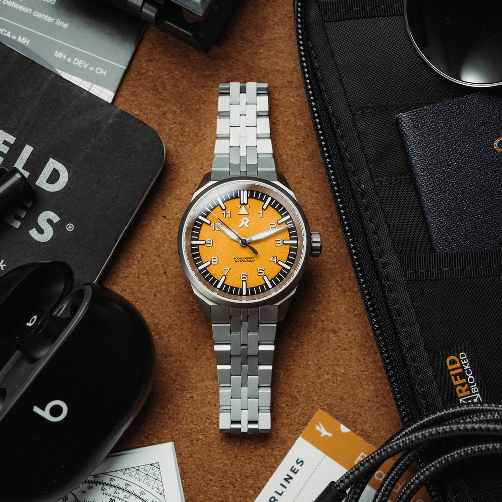 Silver watch with a yellow face on a brown surface with electronic devices and a wallet.